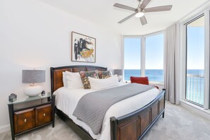 A bedroom with a large bed, a ceiling fan, a nightstand, a lamp, and a window with a view of the ocean.