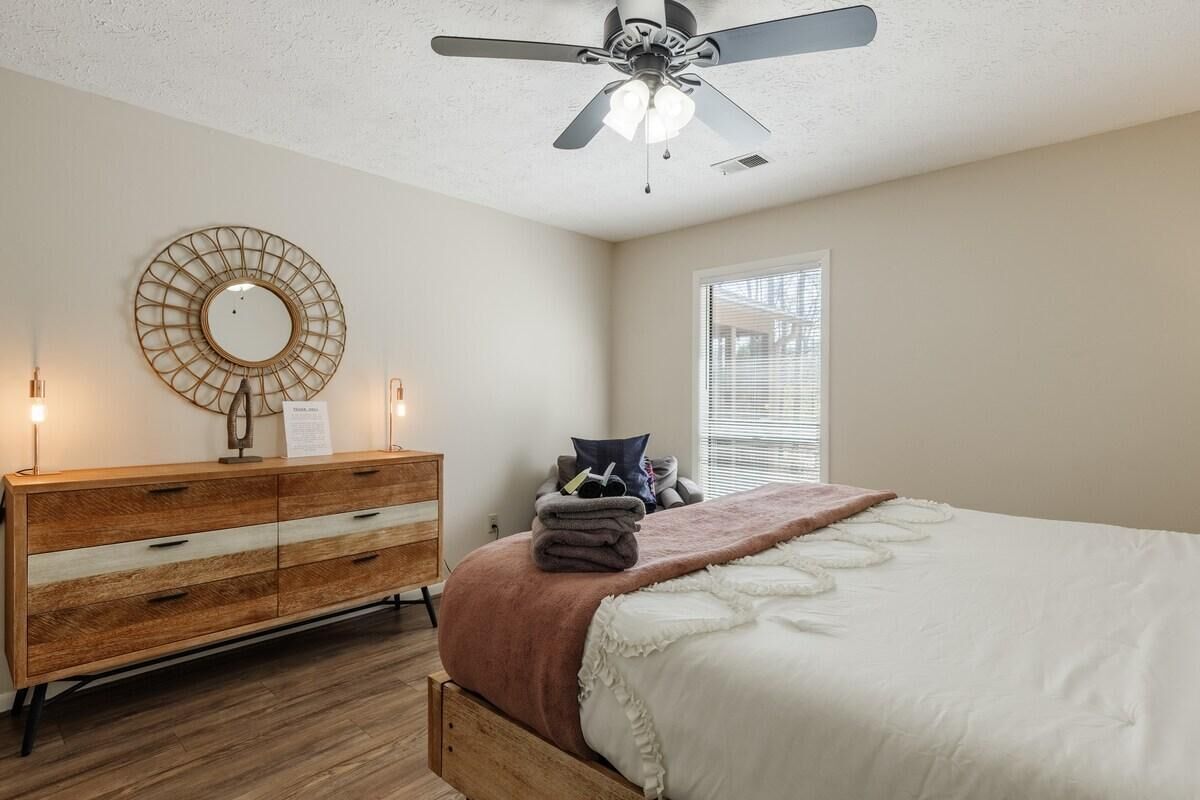 Un dormitorio con cama, cómoda de madera, ventilador de techo, espejo redondo y ventana con persianas.