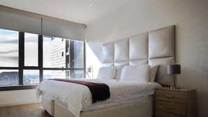 Un dormitorio moderno con una cama grande, una mesita de noche y vistas a la ciudad a través de amplias ventanas.