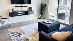 Un salon moderne doté d’un téléviseur à écran plat, de meubles aux couleurs sombres, d’une grande fenêtre et offrant une vue sur un quartier résidentiel.
