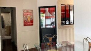 Een eethoek met een glazen tafel en stoelen, een zwarte deur met een rood-wit bord, en een plank met spullen aan de muur.