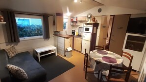 Een knus woongedeelte met een bank, een eettafeltje, een kitchenette en een raam met gordijnen.