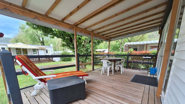 Een overdekt terras buiten met een houten tafel en stoelen, een rieten lounge stoel, en een kleine tafel met een blauwe emmer.