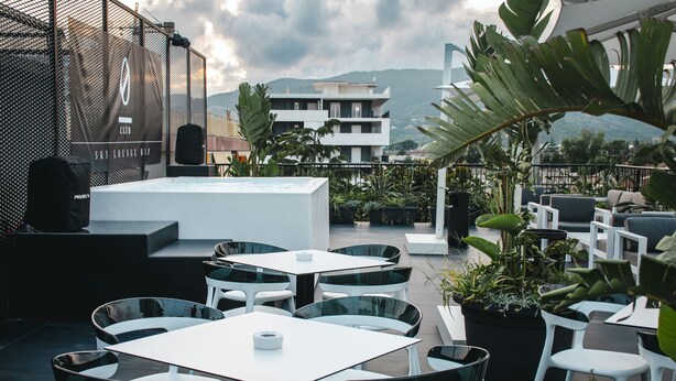 Un lounge sul tetto con tavoli e sedie bianche, circondato da piante in vaso e con vista sulla città.