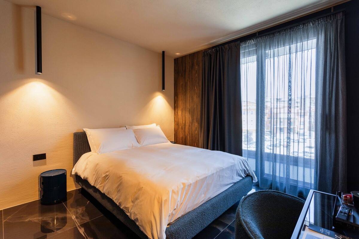 Une chambre d’hôtel moderne dotée d’un grand lit, d’un bureau avec une chaise et offrant une vue sur la ville à travers de fines rideaux.