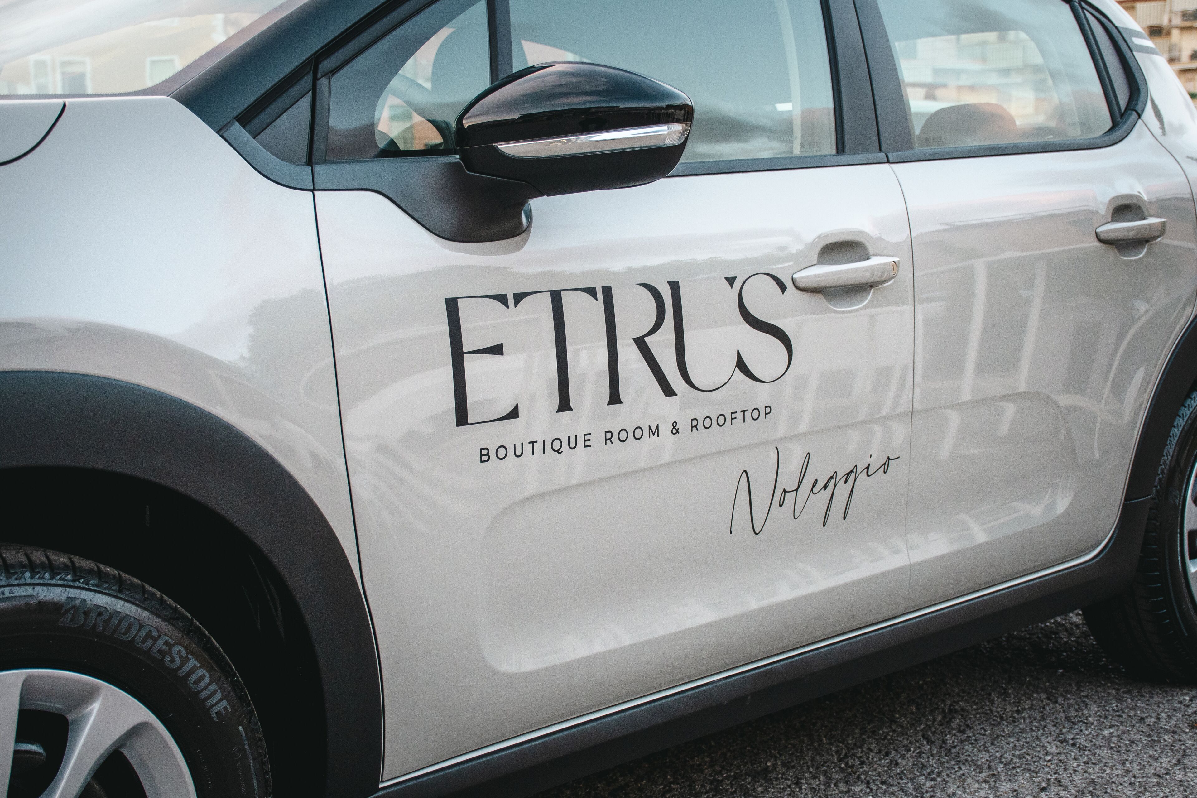 A white car with the text "ETRUS BOUTIQUE ROOM & ROOFTOP" and "Notteggio" on its side.