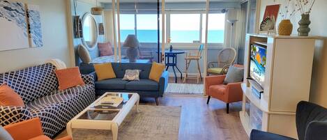 Una sala de estar con sofá, sillones, una mesa de centro y televisor. Se ve el océano a través de amplias ventanas.
