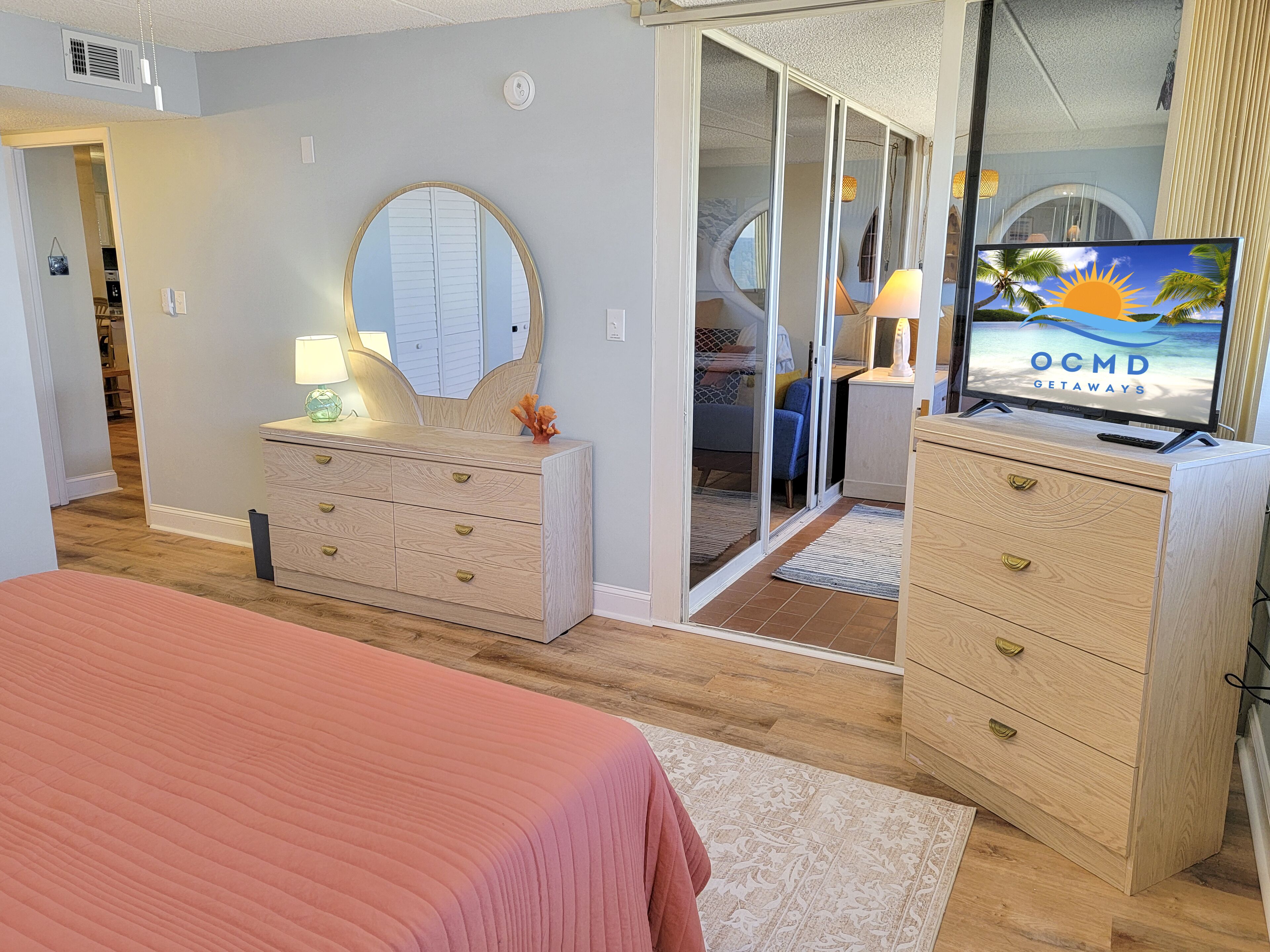Un dormitorio con cama, cómoda, espejo y televisor mostrando 'OCMD GETAWAYS'.