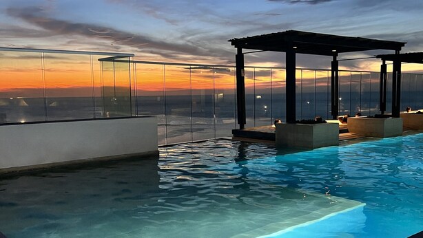 Une piscine à débordement avec une barrière en verre, une pergola et une vue imprenable sur le coucher de soleil sur l’eau.