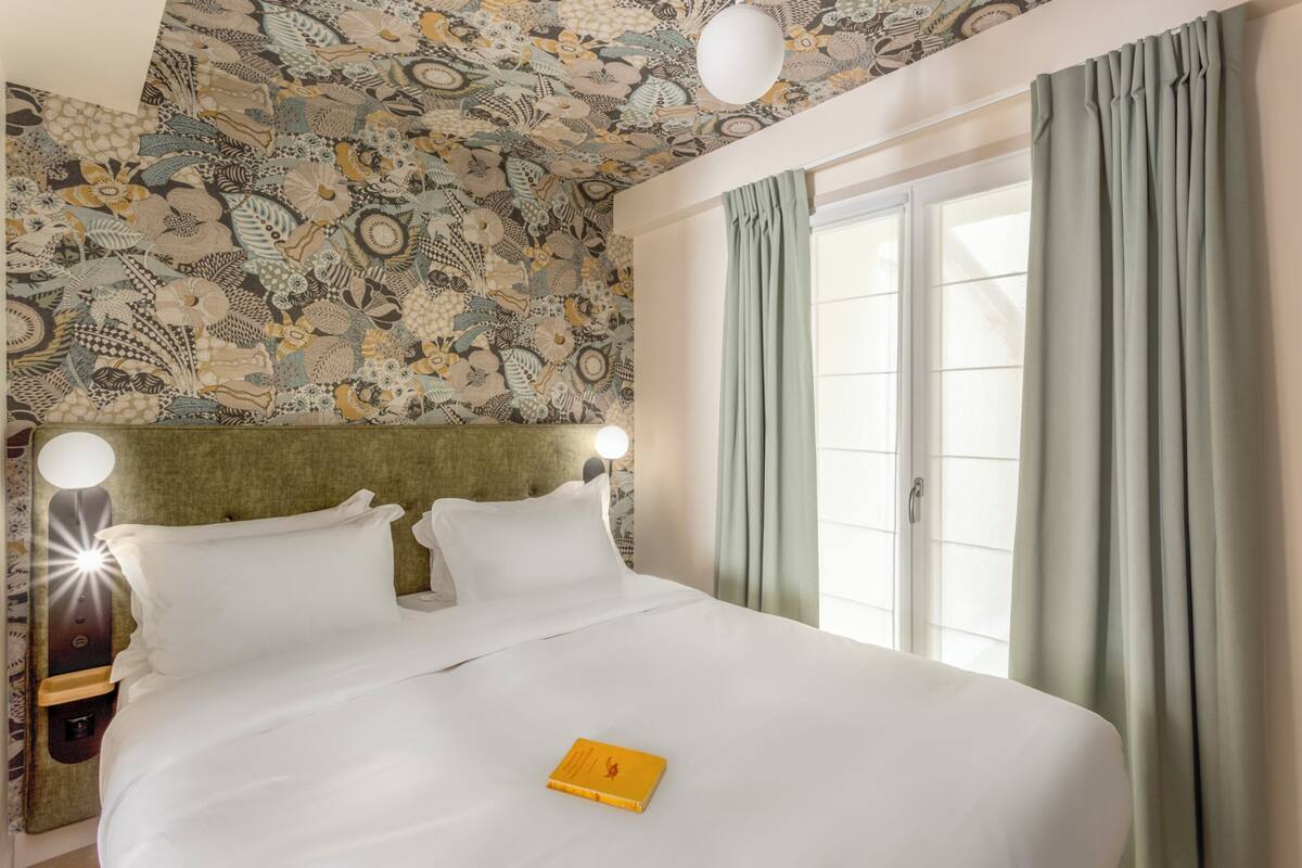 A neatly made bed with white linens, a yellow book on the bed, and a patterned ceiling.
