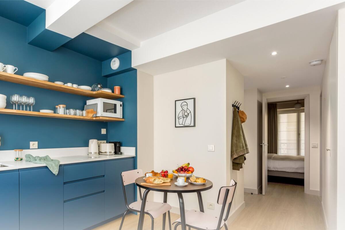 A modern kitchen with blue cabinets, wooden shelves, and a dining area with a table set for breakfast.