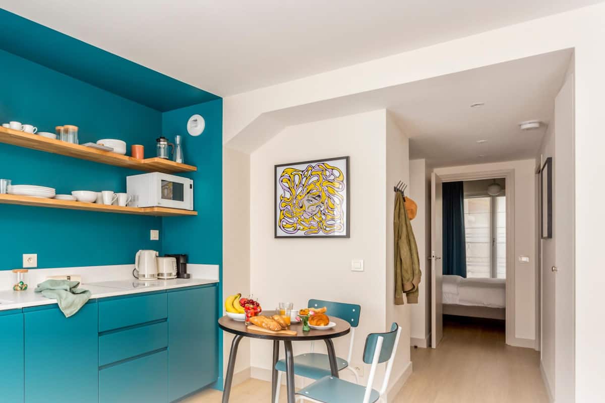 A modern kitchen with teal cabinetry, white countertops, and wooden shelves. There is a dining area with a table set for two, chairs, and a fruit bowl. A microwave and coffee maker are visible on the counter.