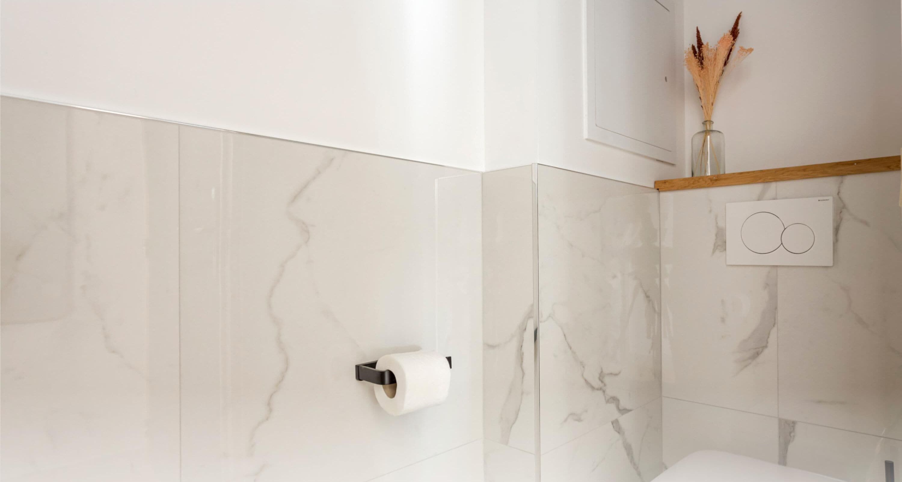 Un baño con paredes de mármol, un dispensador de papel higiénico y un jarrón decorativo con plantas secas.