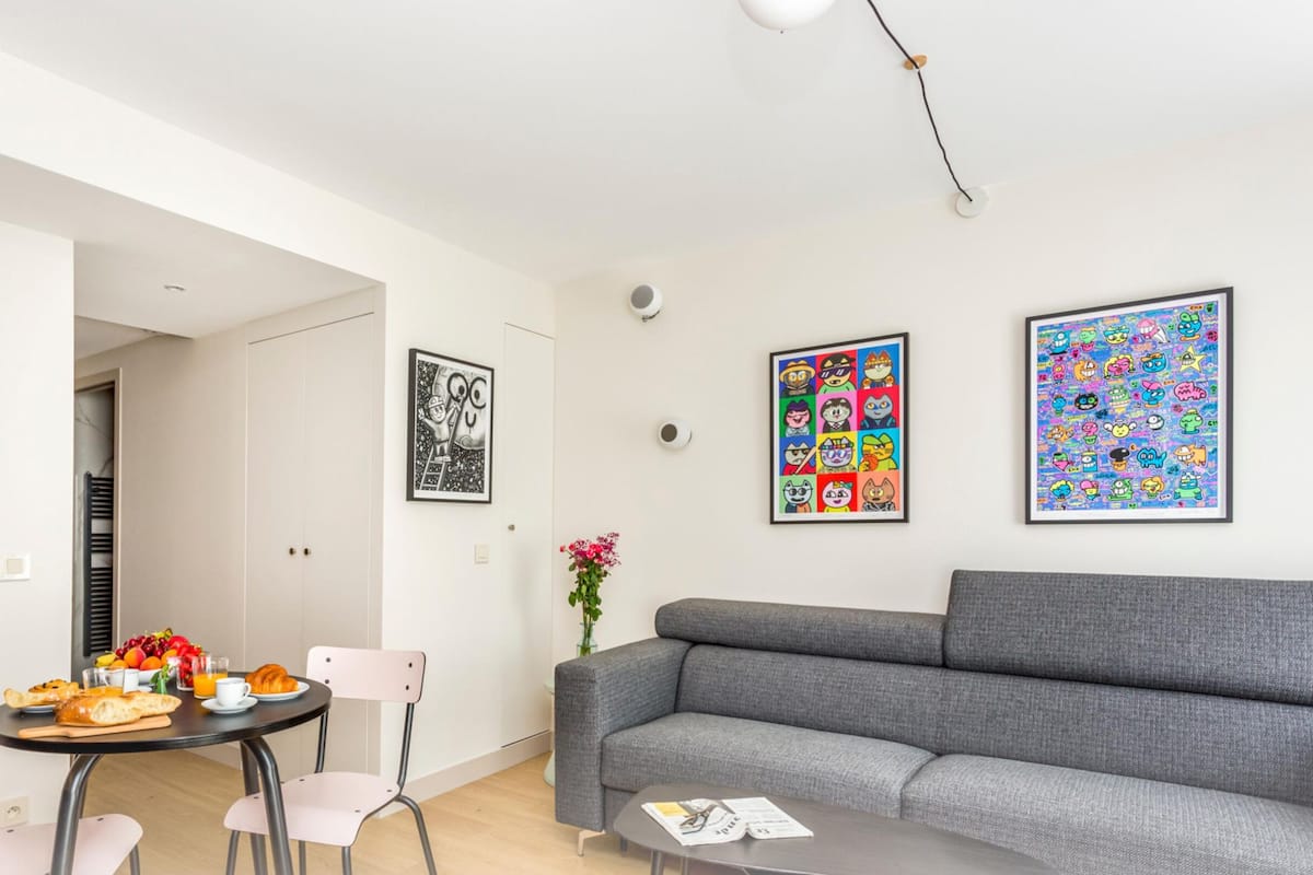 A modern living room with a grey sofa, a dining table with breakfast, and colorful wall art.