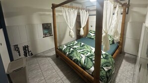 A four-poster bed with a canopy, a single bed with a green leaf-patterned bedspread, a wall-mounted shelf with a picture, and a tiled floor. - Tartane Beach SPA 2 MER (La Trinité)