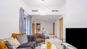 A modern living room with a grey sofa, a coffee table with a vase and remote, a TV, and a dining area in the background. - Tanin - Modern Spacious 1BR Apartment With 2 Balconies (Dubai)