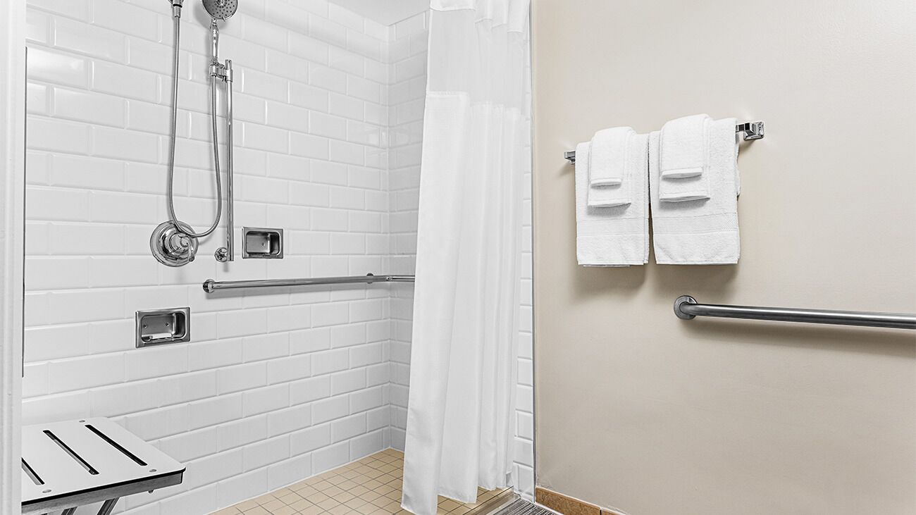 Une salle de bain avec des murs carrelés de carreaux blancs, une douche équipée d’un pommeau de douche à main, d’une barre d’appui et de deux serviettes posées sur un porte-serviettes.
