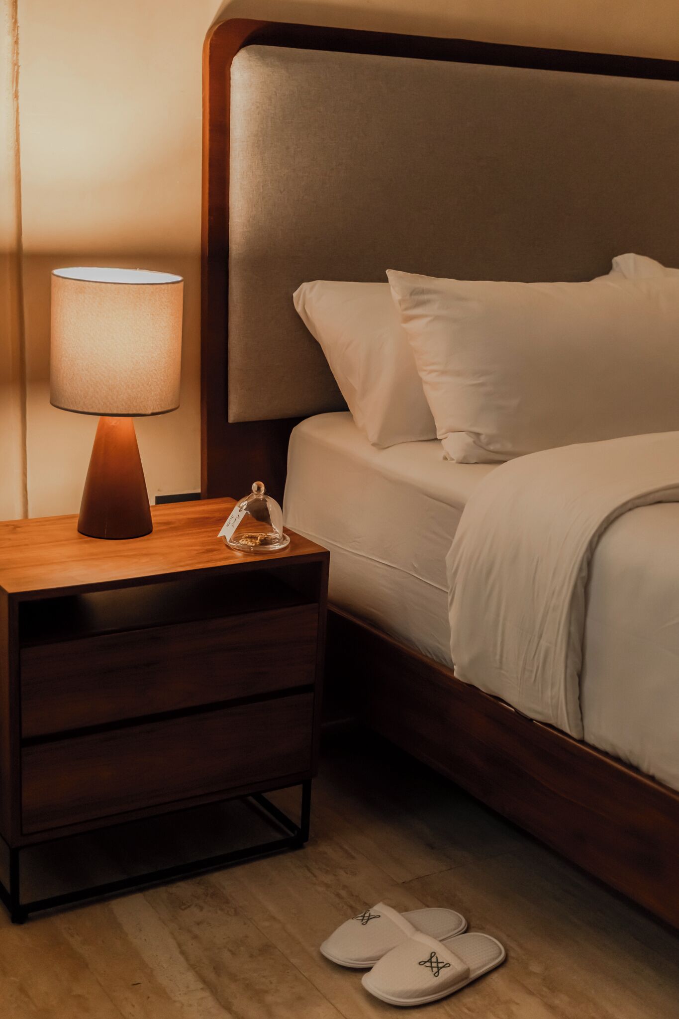 A neatly made bed with white linens, a wooden nightstand with a lamp, and a pair of slippers on the floor.