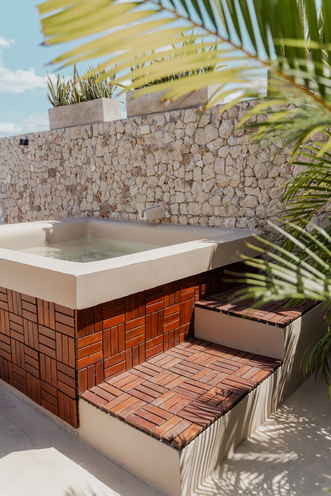 A hot tub with a stone wall backdrop, wooden steps, and potted plants.