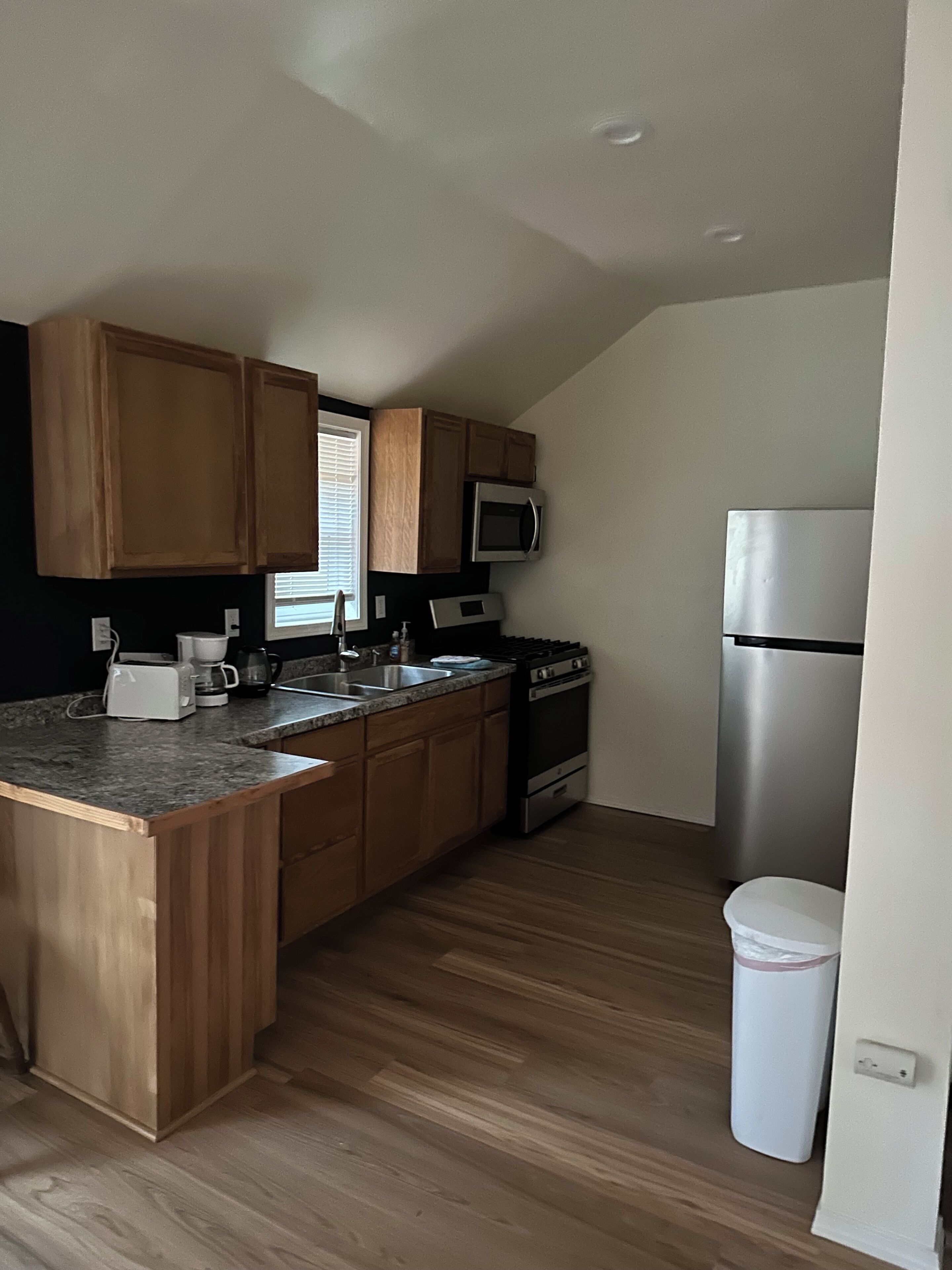 A kitchen with wooden cabinets, a granite countertop, a microwave, a stove, and a trash can.