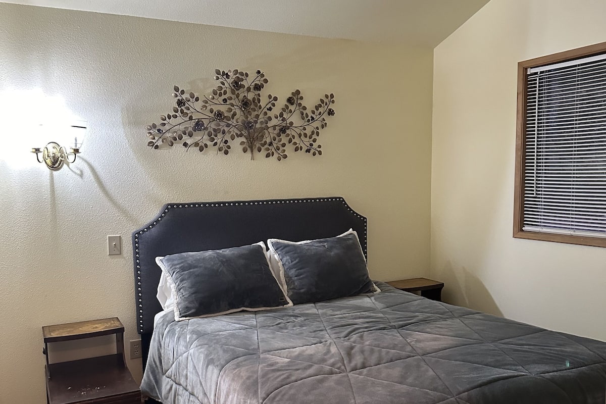 A bedroom with a bed, two nightstands, a wall-mounted light fixture, and a window with blinds.