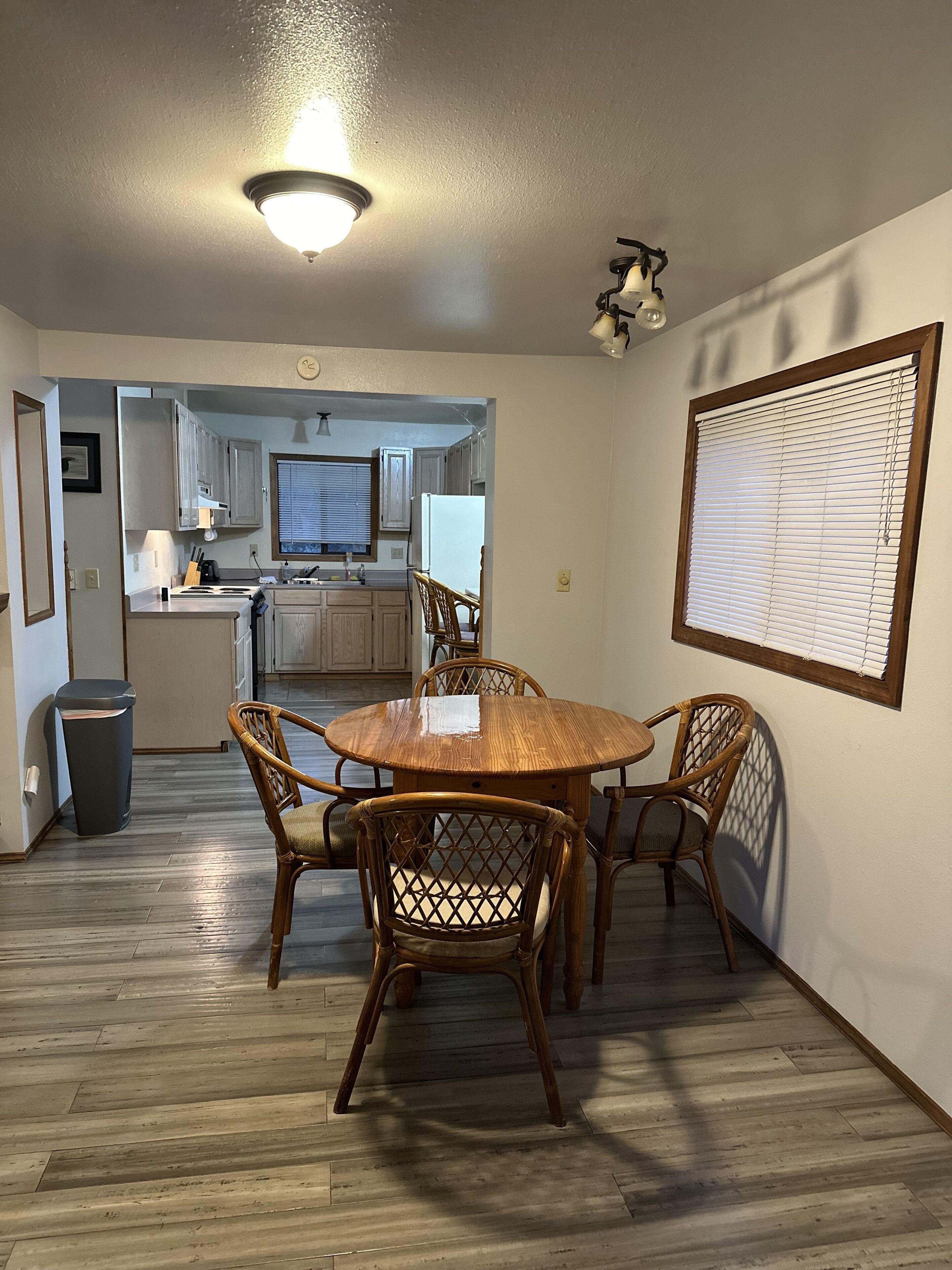 A dining area with a wooden table and chairs, a kitchen in the background, and a window with blinds.