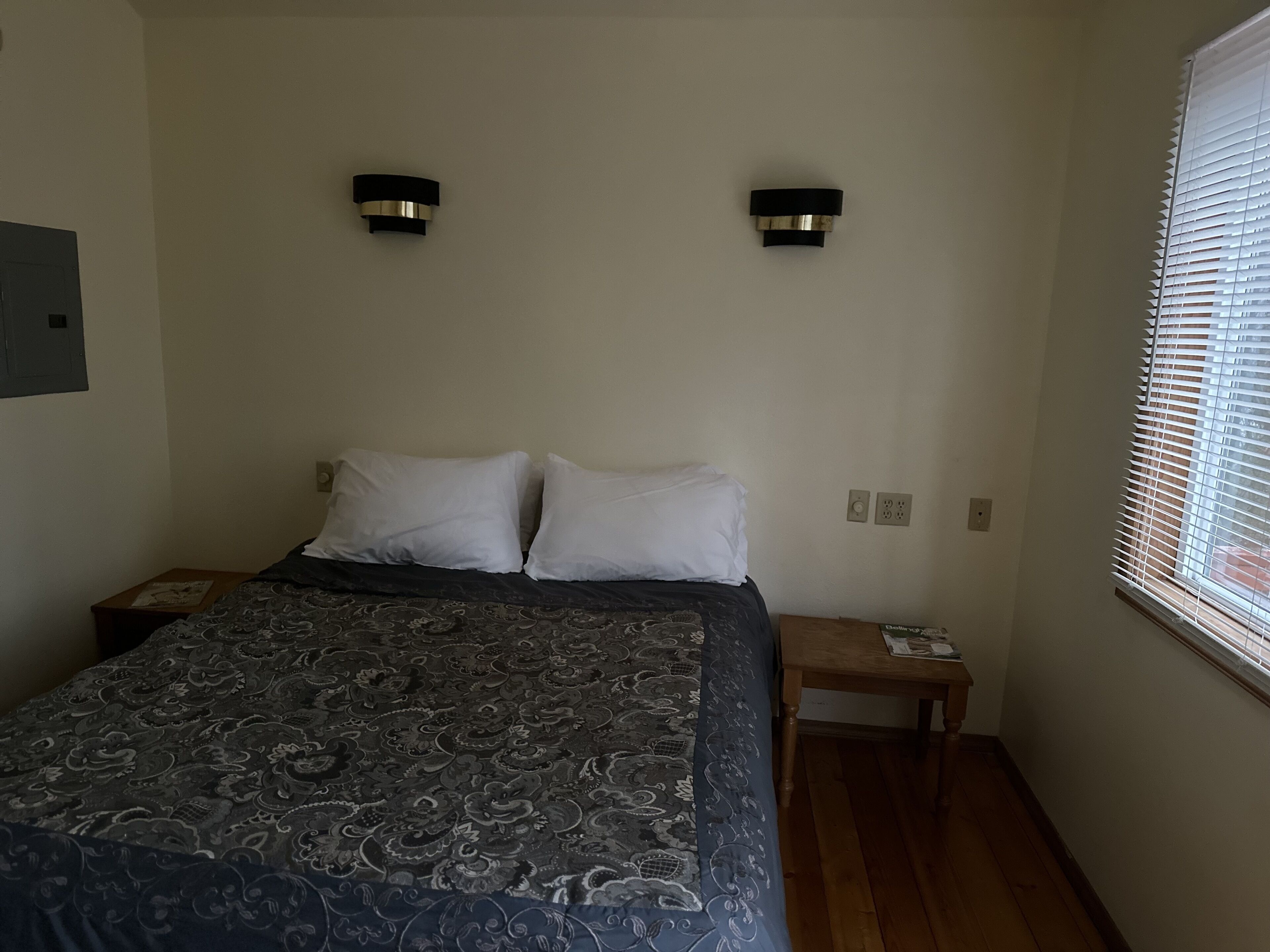 A bedroom with a bed, two bedside tables, a nightstand, a window with blinds, and two wall-mounted lights.