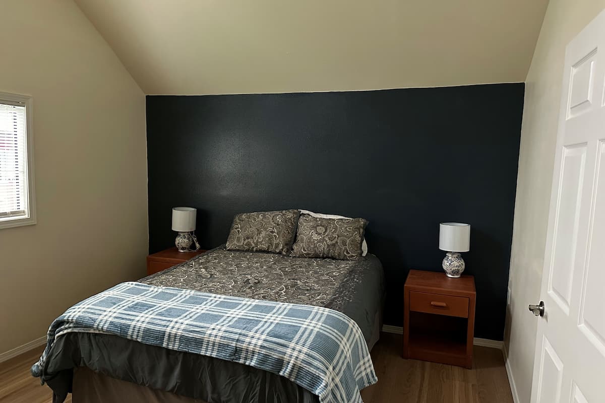 A bedroom with a dark accent wall, a bed with patterned bedding, two bedside tables with lamps, and a window with blinds.