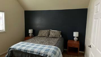 A bedroom with a dark accent wall, a bed with patterned bedding, two bedside tables with lamps, and a window with blinds.