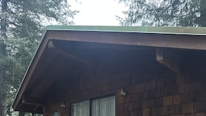 Una terraza de madera con un banco y una mesa, una escalera que lleva a un área cubierta con una puerta corrediza de vidrio, y una parrilla de barbacoa al fondo.