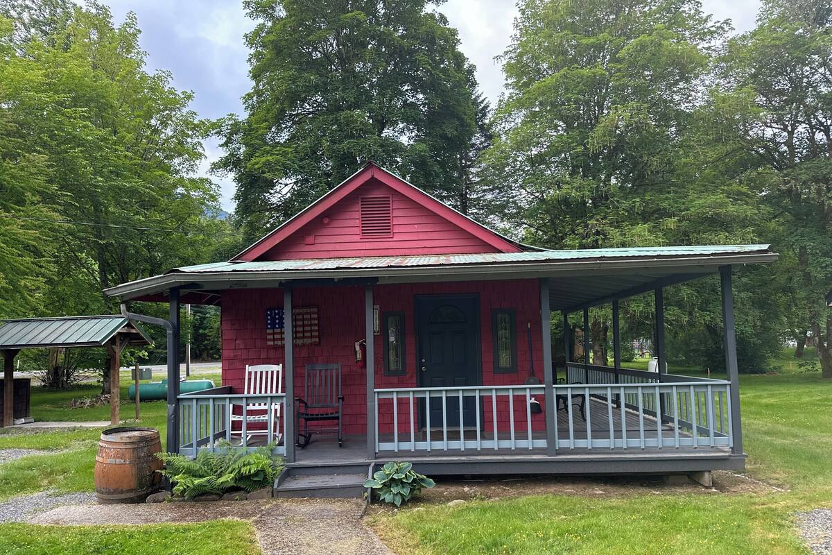 A red wooden cabin with a porch, a flag, and a small garden area.