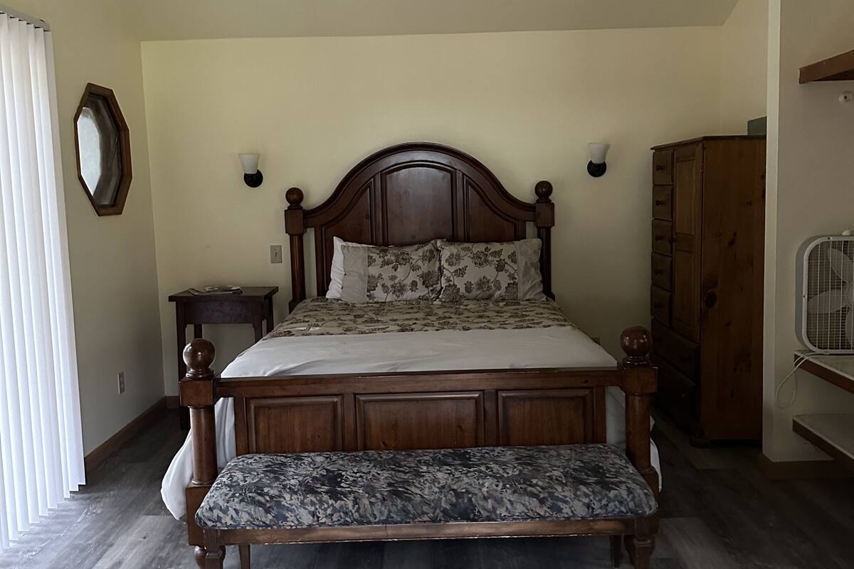 A bedroom with a wooden bed, a bench, a ceiling fan, a wooden wardrobe, and a window with blinds.