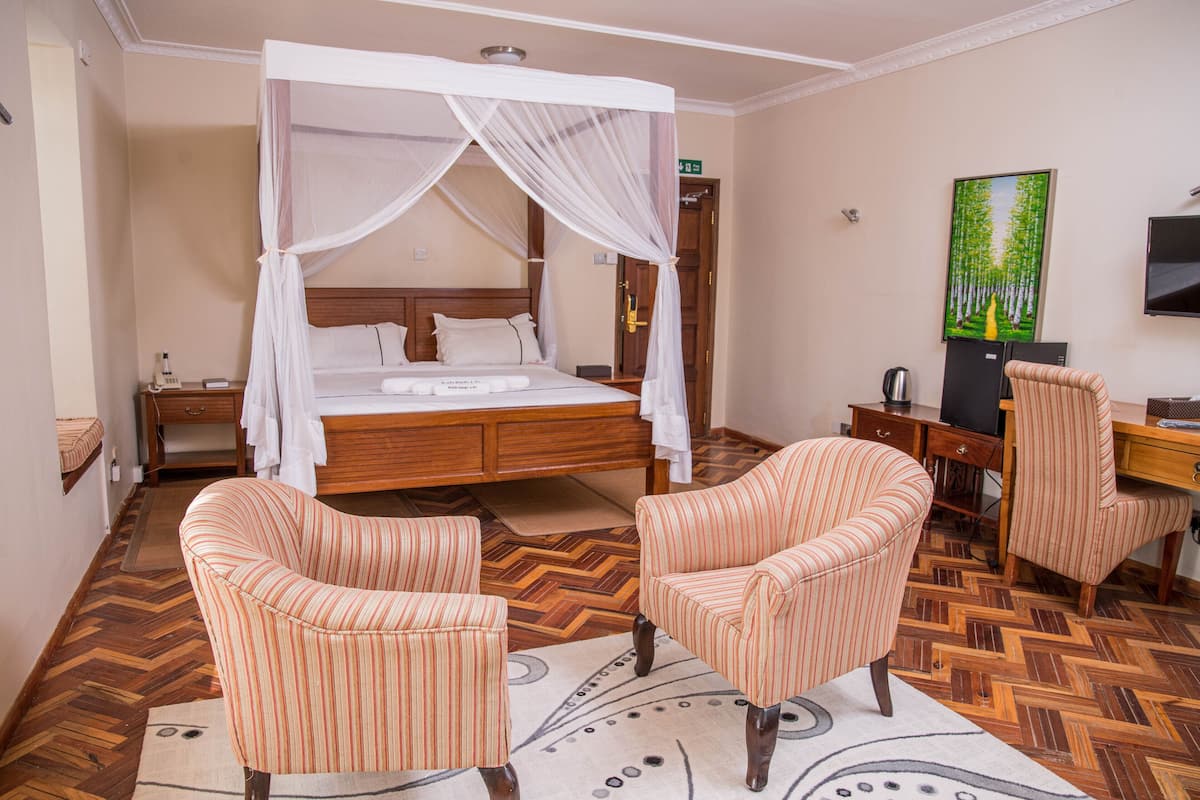 A hotel room with a four-poster bed, two striped armchairs, a desk with a chair, a television, and a painting on the wall.