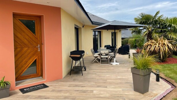 Una casa con puerta de madera, un patio con mobiliario exterior y un jardín con plantas.