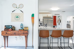 A modern kitchen with white cabinets, a stainless steel refrigerator, and a red accent wall. There are three brown bar stools and a wooden table with vintage items. - Family Fun on Guadalupe River w/ kayaks, grill and dock! Minutes from Downtown (Seguin)