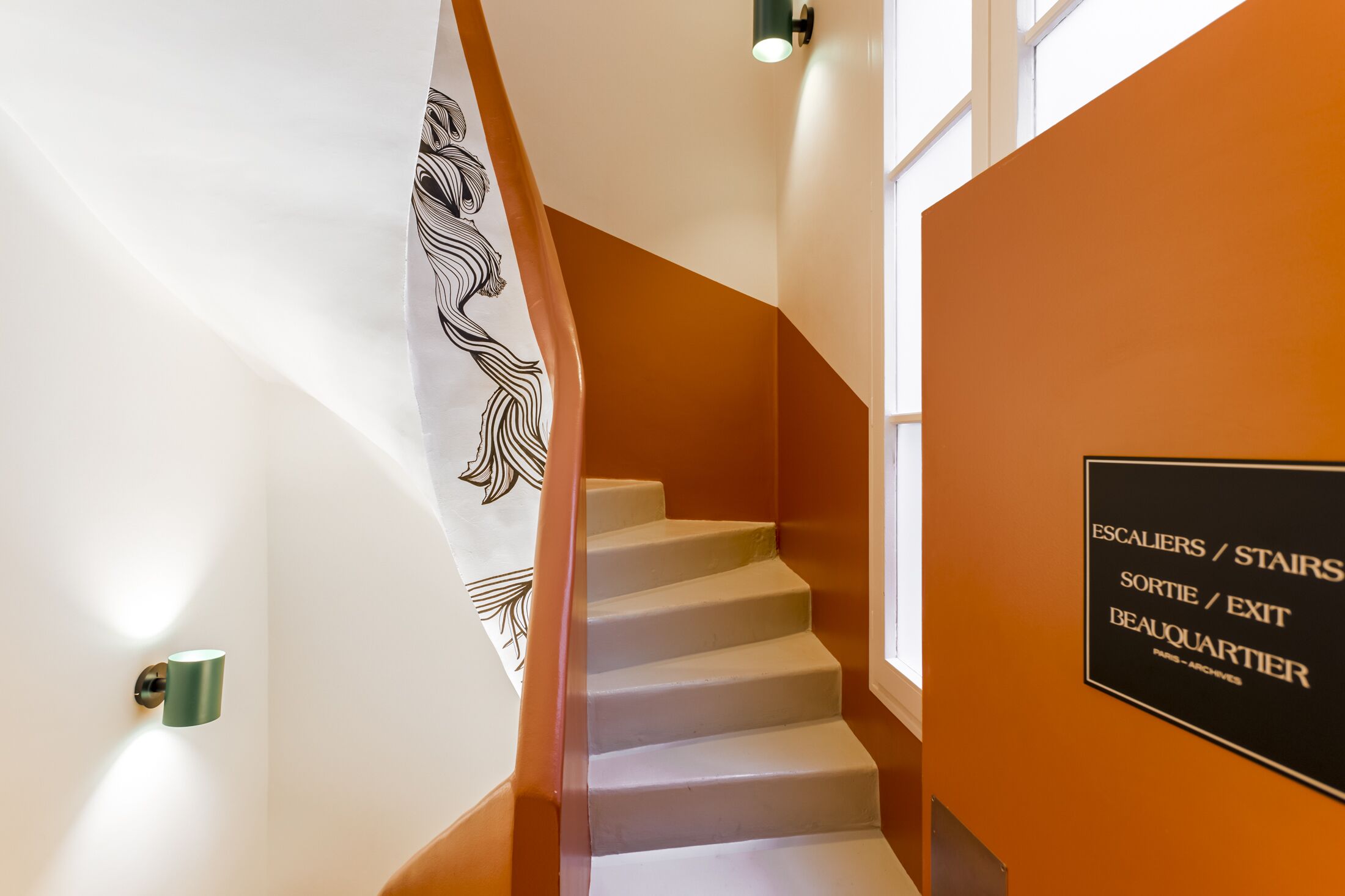 Eine moderne Treppe, ein an der Wand befestigtes Schild mit der Wegbeschreibung und eine Wandleuchte.