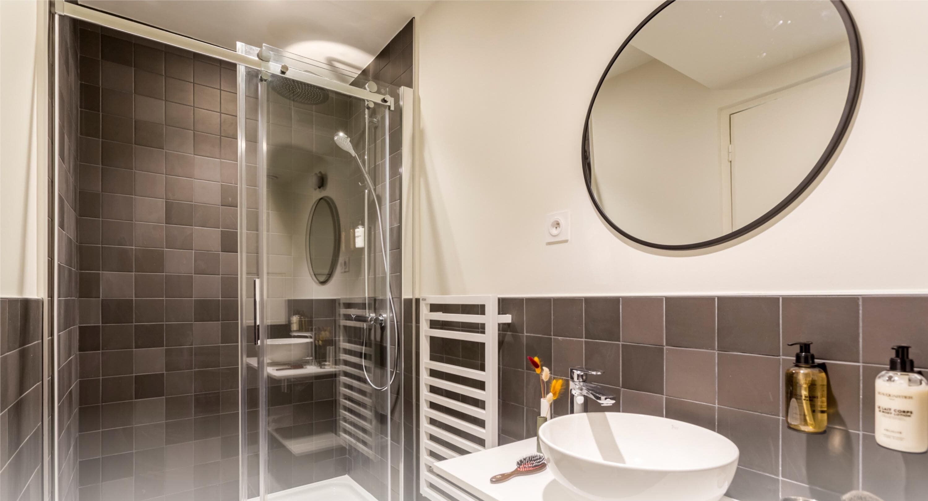 A modern bathroom with a glass-enclosed shower, a white sink, and a round mirror.