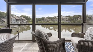 A covered outdoor area with wicker furniture, a view of a lake, and residential buildings in the background.