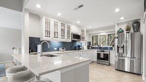 A modern kitchen with white cabinetry, a large island with bar stools, stainless steel appliances, and a blue-tiled backsplash.