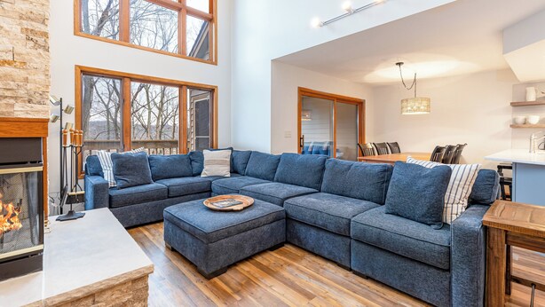 A modern living room with a sectional sofa, a fireplace, large windows, and a kitchen area. - Rock Cove Villa | 3 Kings + Private Baths | Walk to Golf · Fireplace · Foosball (Galena)