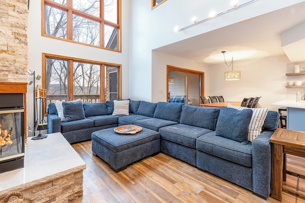 A modern living room with a sectional sofa, a fireplace, large windows, and a kitchen area.