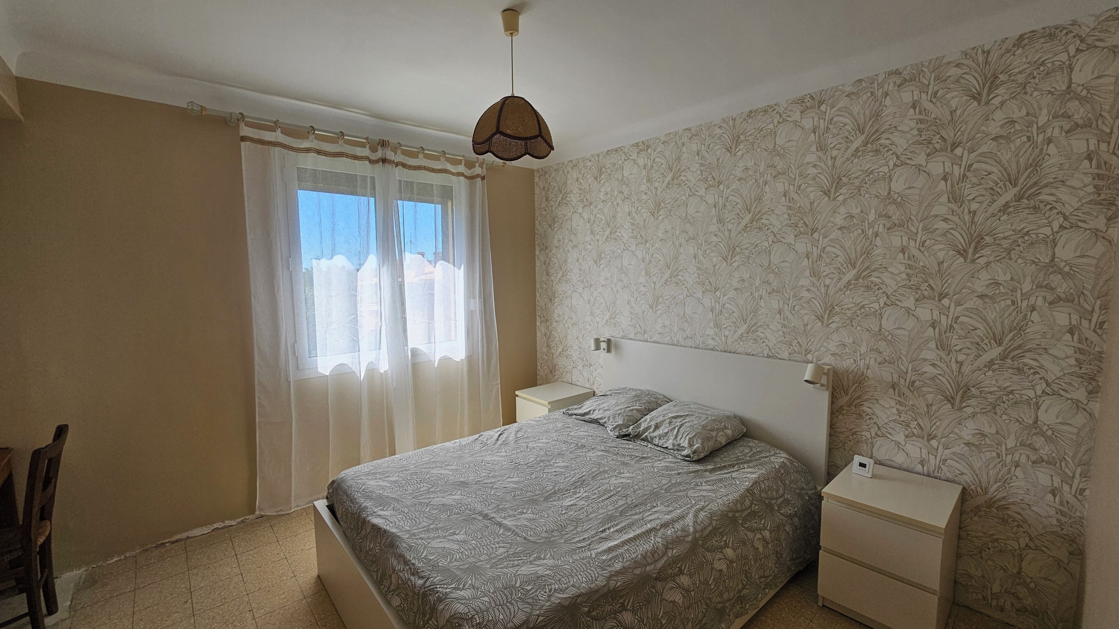 Une chambre à coucher avec un lit, des tables de chevet, une chaise, une fenêtre avec des rideaux et un papier peint à motifs.