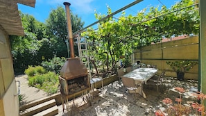 A patio with a brick floor, a table with chairs, a pizza oven, and a garden with trees and plants. - Maison Entre mer et Montagne (Saint-Estève)