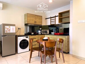 A compact kitchen with wooden cabinets, a washing machine, a microwave, and a dining area with wooden chairs. - Timurbay Seafront Residences NatureHome (Kuantan)