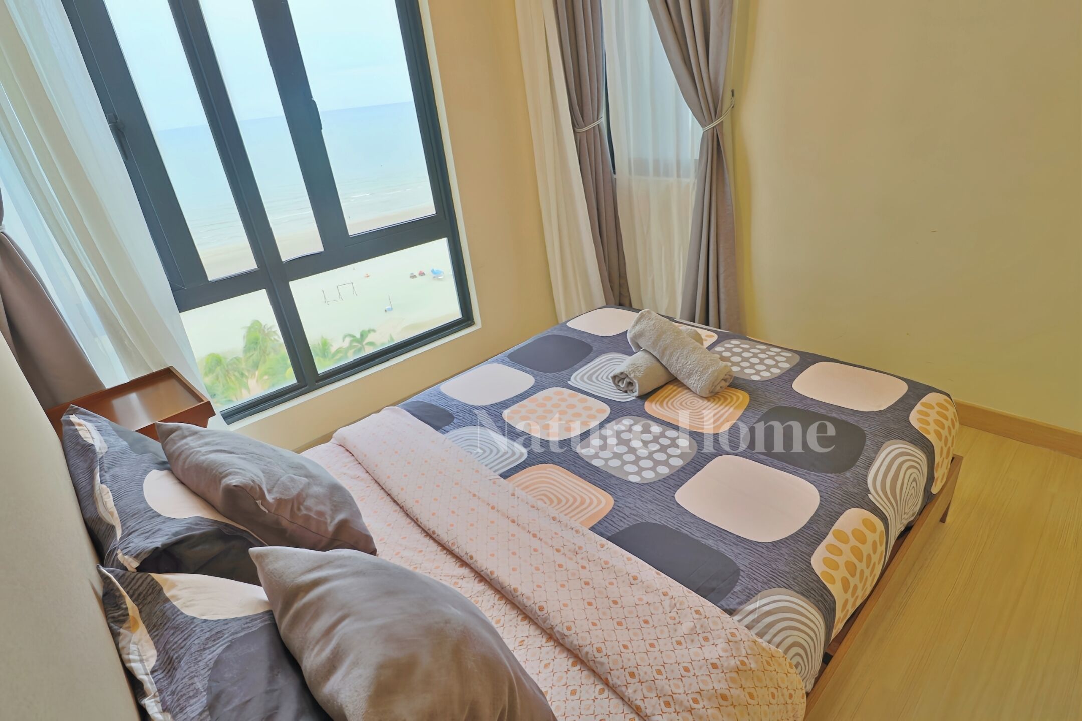 A bed with a patterned bedspread, pillows, and a view of the beach through a window.