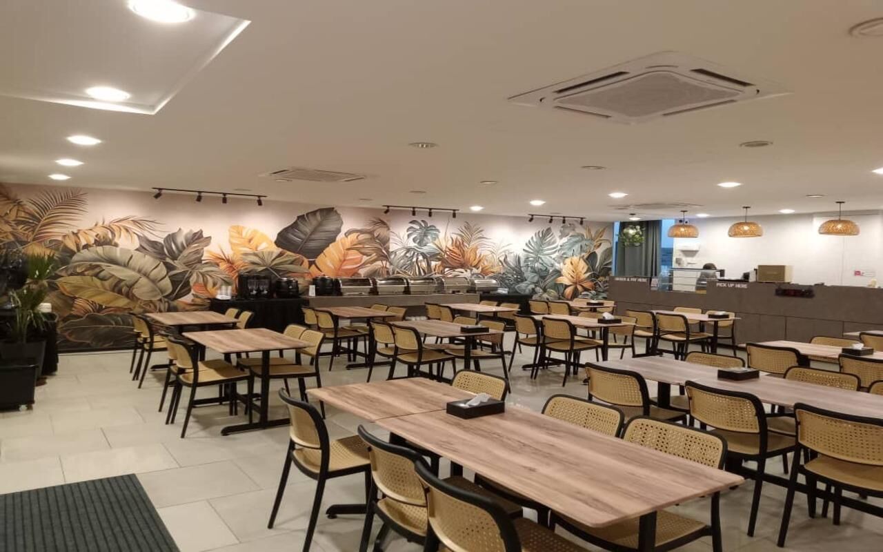 Um restaurante com várias mesas e cadeiras, um mural com folhas tropicais na parede, e um balcão com uma placa que diz 'Retirar Pedidos'.