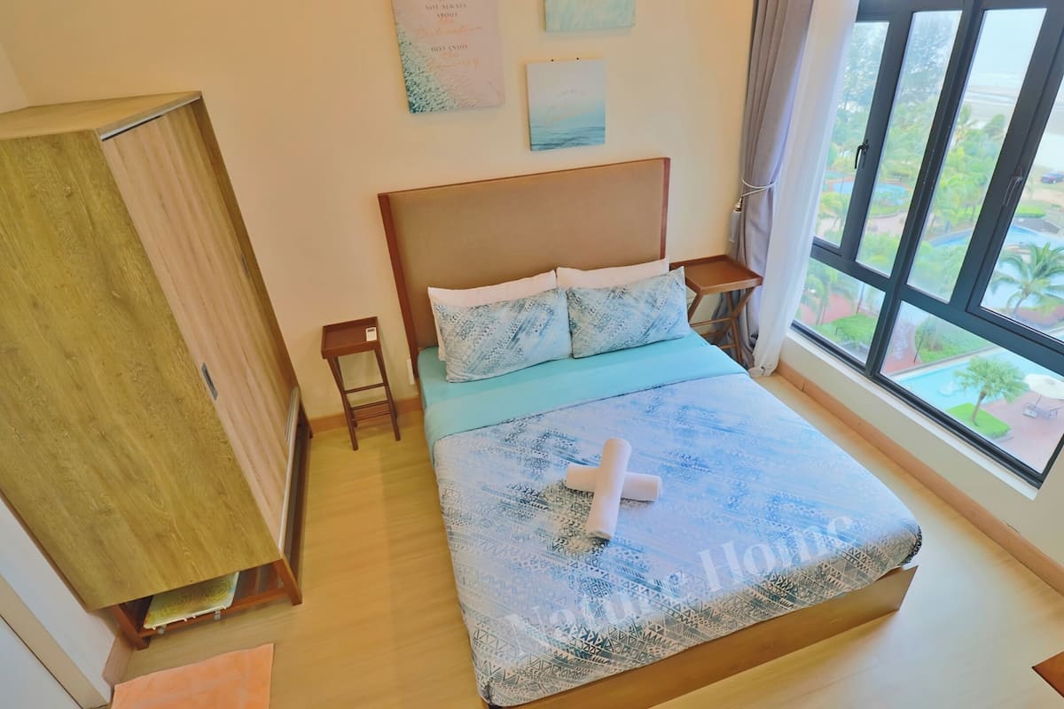 A bedroom with a wooden headboard, a patterned bedspread, and a view of a pool and greenery through a large window.