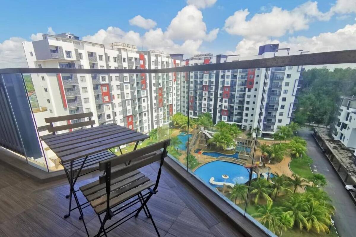 A balcony with a wooden table and chairs, overlooking a residential area with modern buildings and a swimming pool.