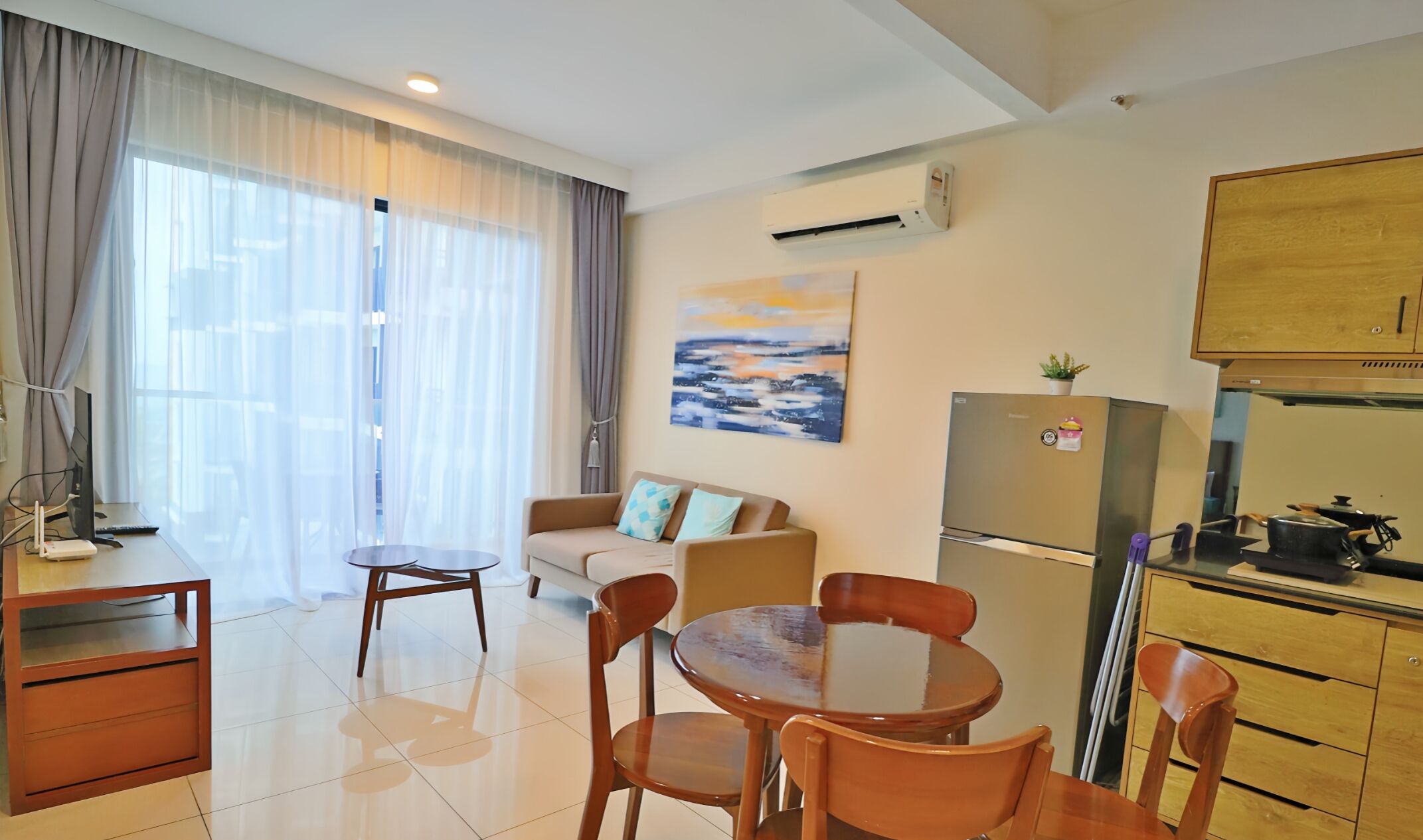 Una sala moderna con sofá, una mesa de comedor con sillas, una zona de cocina con refrigerador y estufa, y ventanas amplias con cortinas livianas.