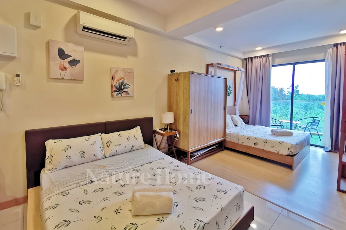 A hotel room with two beds, a wardrobe, a bedside table with a lamp, and a view of greenery through the window.